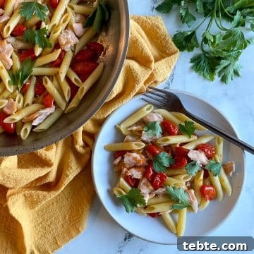 A white plate of penne pasta with burst cherry tomatoes and flaked salmon.