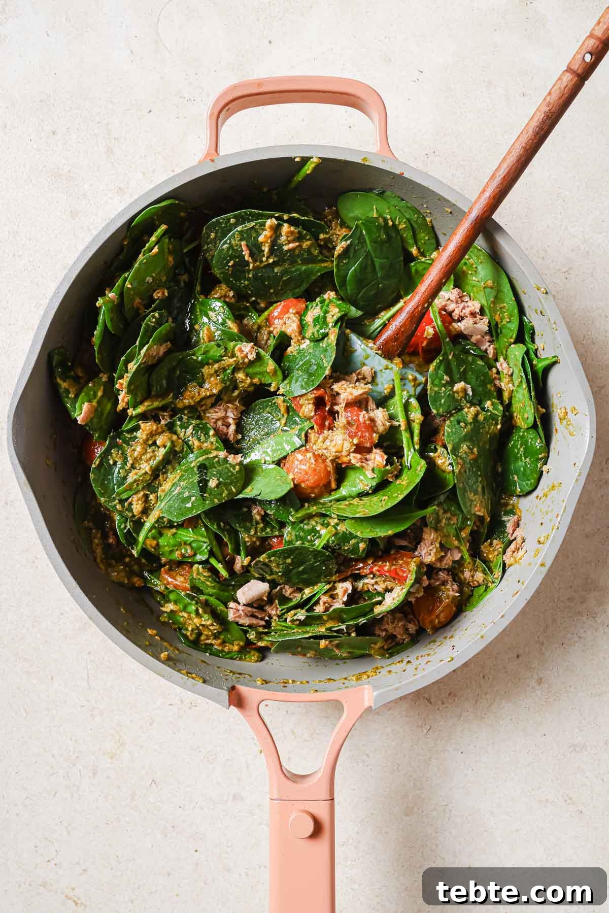 Creamy Tuna Pesto Pasta Recipe for Quick Weeknight Meals 8 A skillet filled with spinach, tomatoes, and tuna with a wooden spoon.