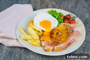 Classic Gammon Steak with Fried Egg and Chips 9 Gammon steak served with homemade chips, duck egg with the yolk pouring over the ham, peas, vined cherry tomatoes and two pineapple rings on a white plate