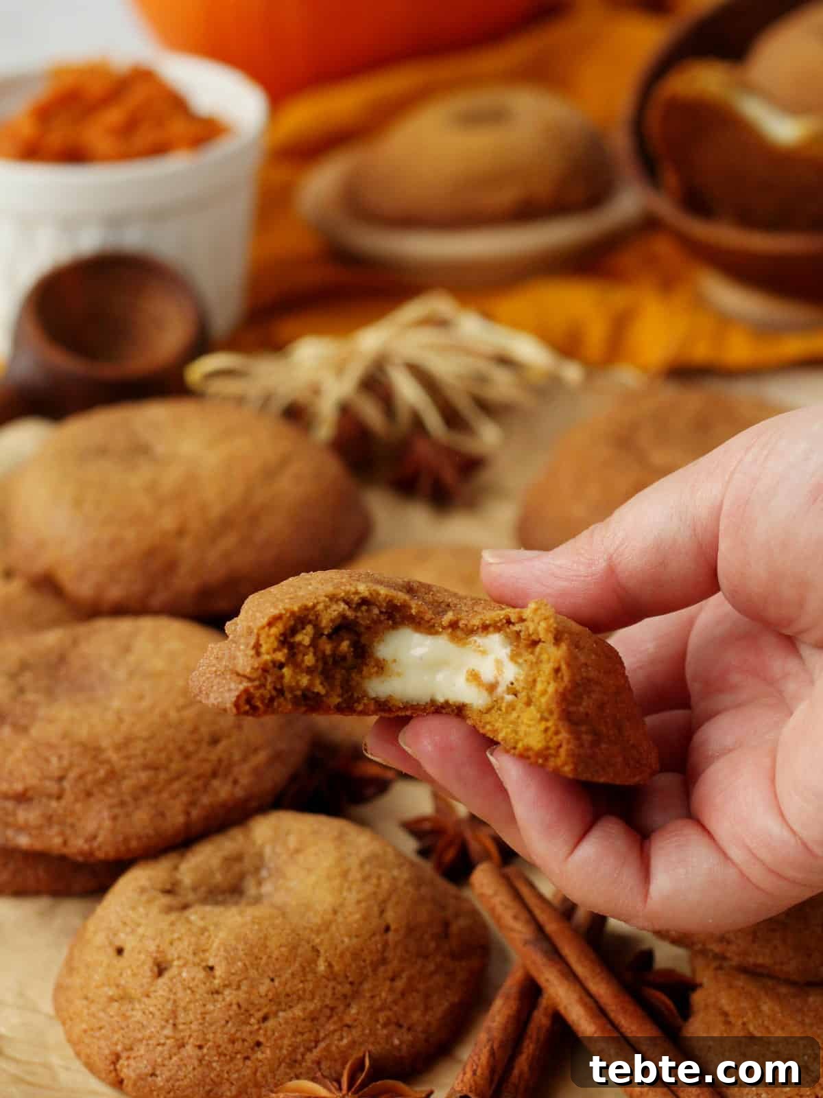 Pumpkin Cheesecake Cookies Recipe — Soft, Creamy Fall Treat 2 pumpkin cheesecake cookies placed on parchmend paper with one cookie filled with cream cheese, in the background other cookies and white bowl with pumpkin puree