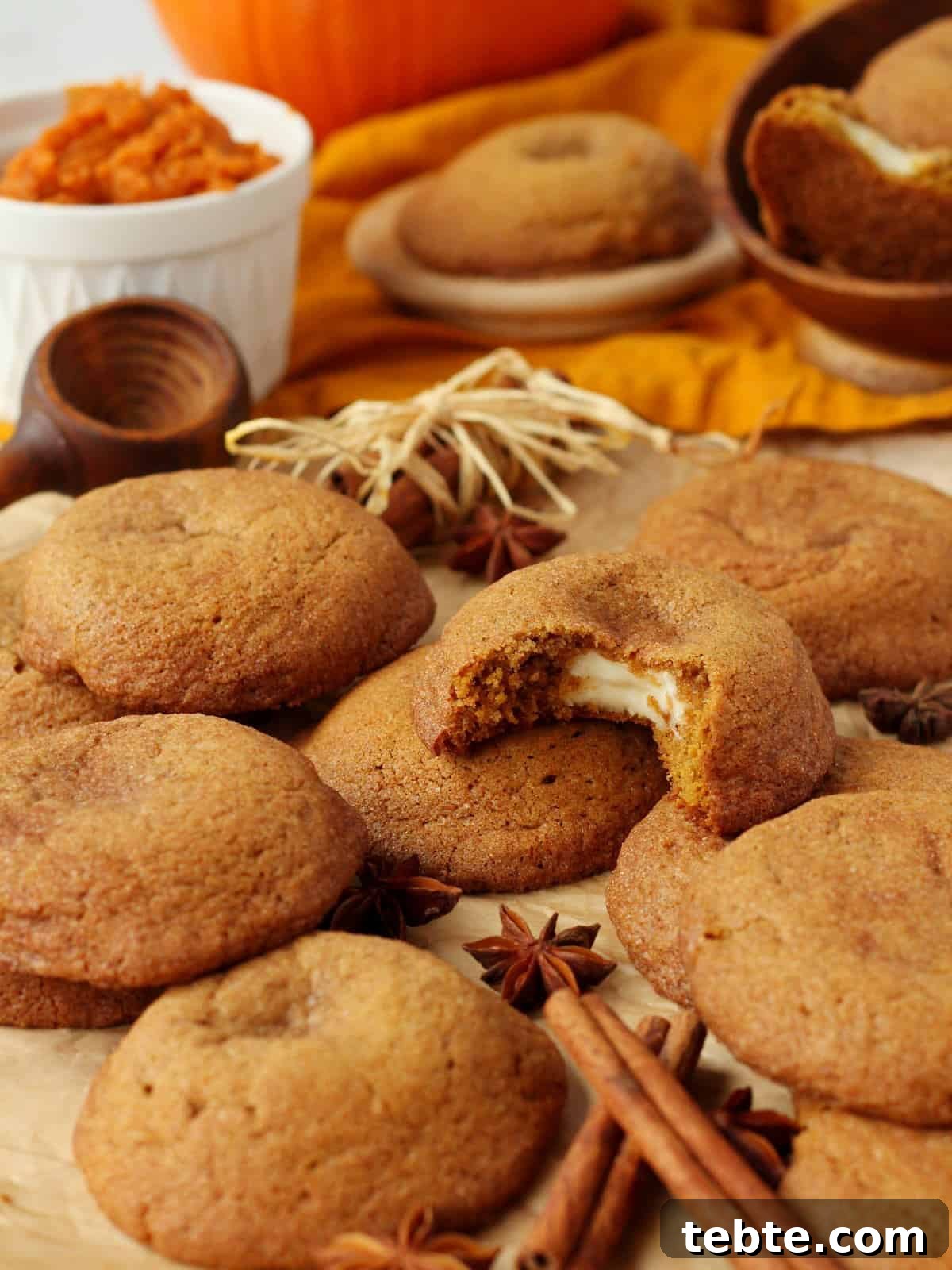 Pumpkin Cheesecake Cookies Recipe — Soft, Creamy Fall Treat 17 Pumpkin cheesecake cookies scattered on brown parchment paper with pumpkin pure & cinnamon sticks in background