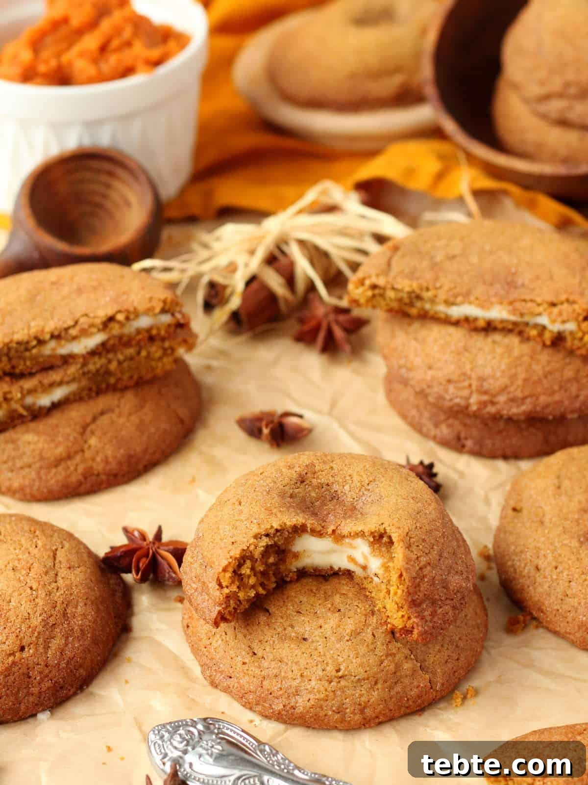 Pumpkin Cheesecake Cookies Recipe — Soft, Creamy Fall Treat 4 warm pumpkin cookies filled with cream cheese, scattered on a brown parchment paper with pumpkin puree and fall decoration in the background