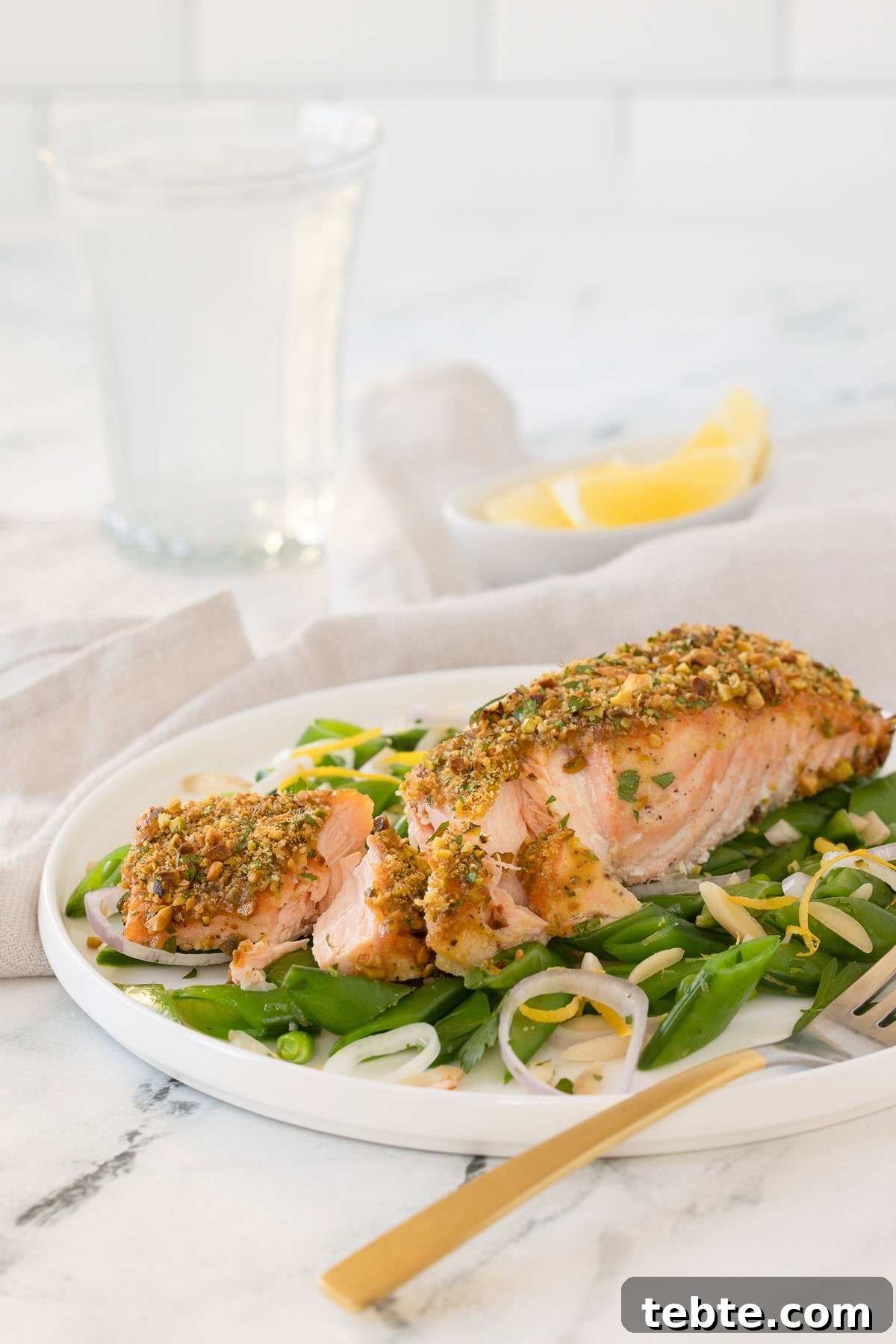 Oven-Roasted Salmon with Pistachio Crust 7 A filet of pistachio-crusted salmon with sugar snap peas and lemon edges in the back, on a white plate.