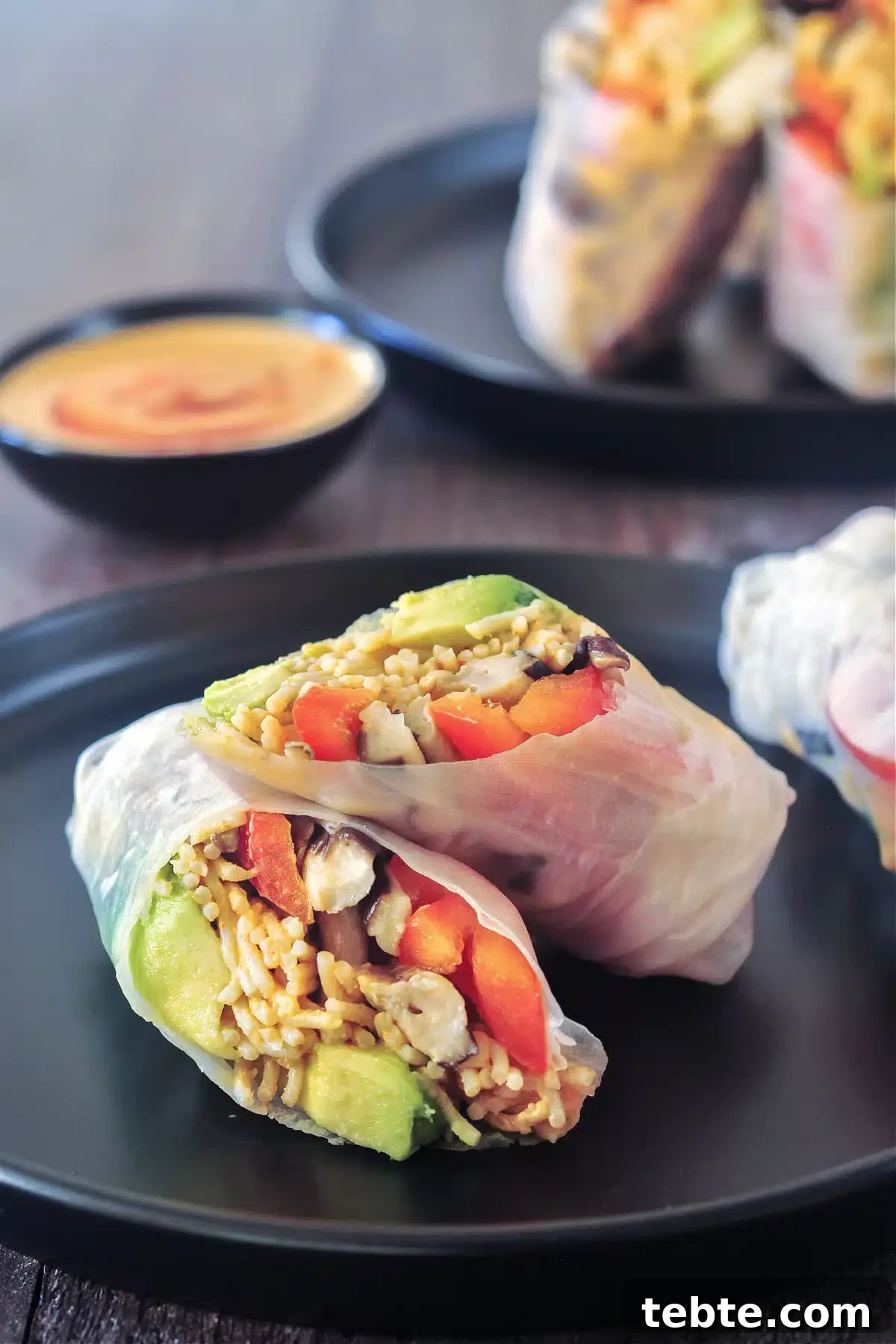 Avocado Shiitake Spring Rolls @spabettie #glutenfree #vegan #fresh #springroll #recipe Two avocado shiitake spring rolls on a matte black plate; one roll is sliced in two pieces to show filling: avocado, shiitake mushrooms, ramen noodles, bell pepper slices, chopped cashews.