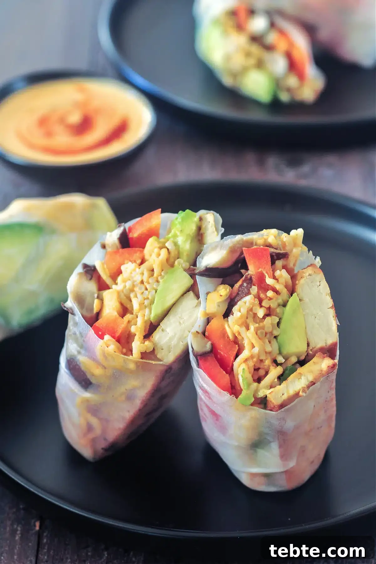 Avocado Shiitake Spring Rolls @spabettie #glutenfree #vegan #fresh #springroll #recipe Two matte black plates of avocado shiitake spring rolls; one roll is sliced in two to show filling: avocado, shiitake mushrooms, ramen noodles, bell pepper slices, chopped cashews. A small dish of spicy almond sauce sits to the side of the plates.