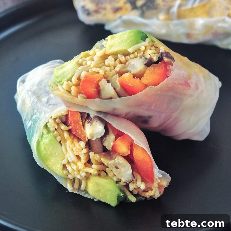 Close up of an avocado shiitake spring roll on a matte black plate; roll is sliced in half to show filling: avocado, shiitake mushrooms, ramen noodles, bell pepper slices, chopped cashews.