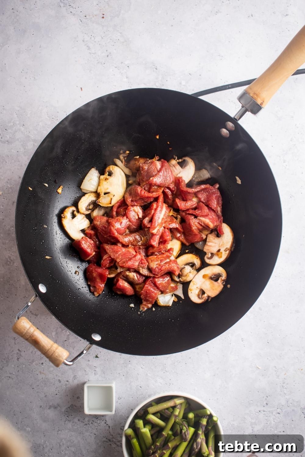 Panda Express Black Pepper Angus Steak Copycat Recipe 7 An overhead view of a hot wok cooking steak and mushrooms. A small bowl of chopped asparagus sits next to it.