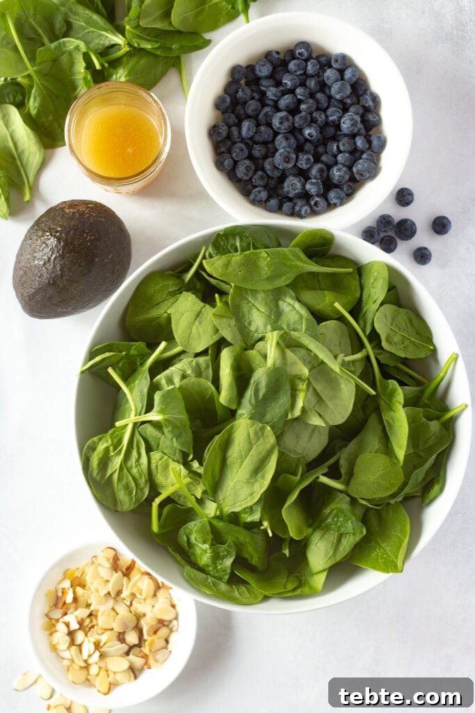 10-Minute Blueberry & Spinach Salad with Sweet Honey Vinaigrette 3 Top down shot of baby spinach, blueberries, sliced almonds, all in white bowls, next to a mason jar of dressing and a whole avocado, all on a white background.