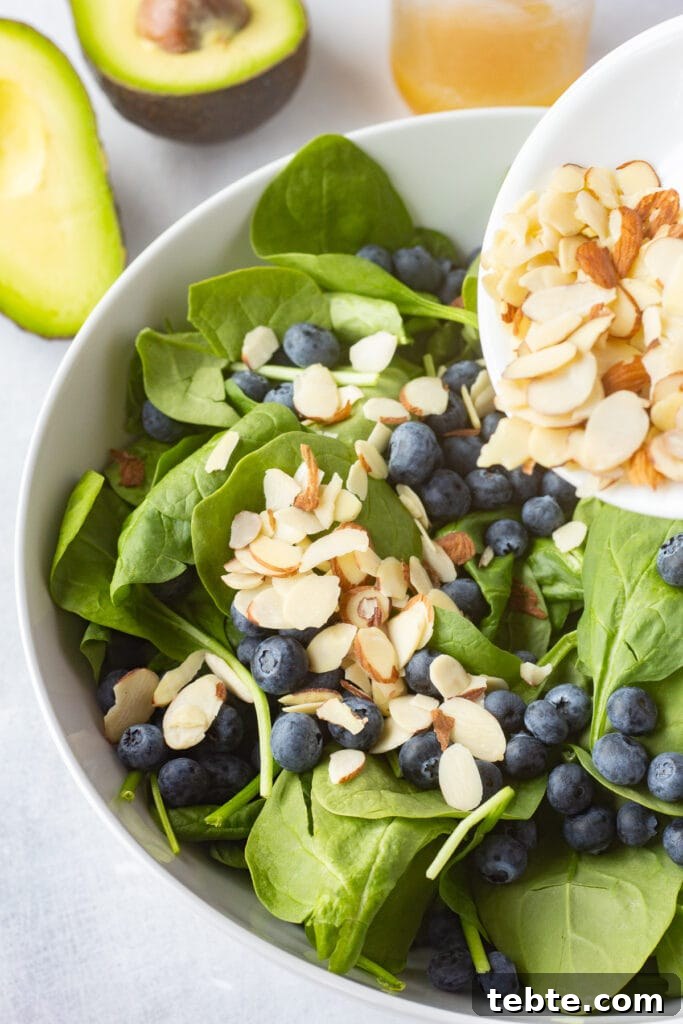 10-Minute Blueberry & Spinach Salad with Sweet Honey Vinaigrette 6 Pouring sliced almonds into a large white salad bowl with baby spinach and fresh blueberries already in it.
