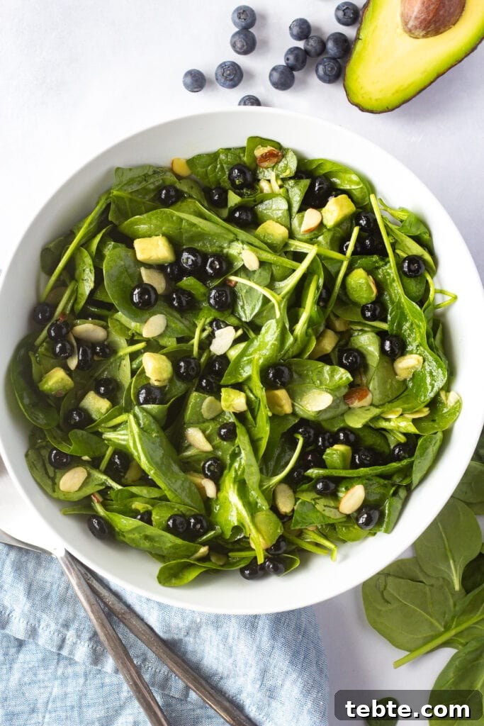 10-Minute Blueberry & Spinach Salad with Sweet Honey Vinaigrette 9 Top down shot of a large white serving bowl with blueberry spinach salad in it. A blue napkin and serving utensils are in the bottom left corner and ingredients for the salad are in other corners of the frame.