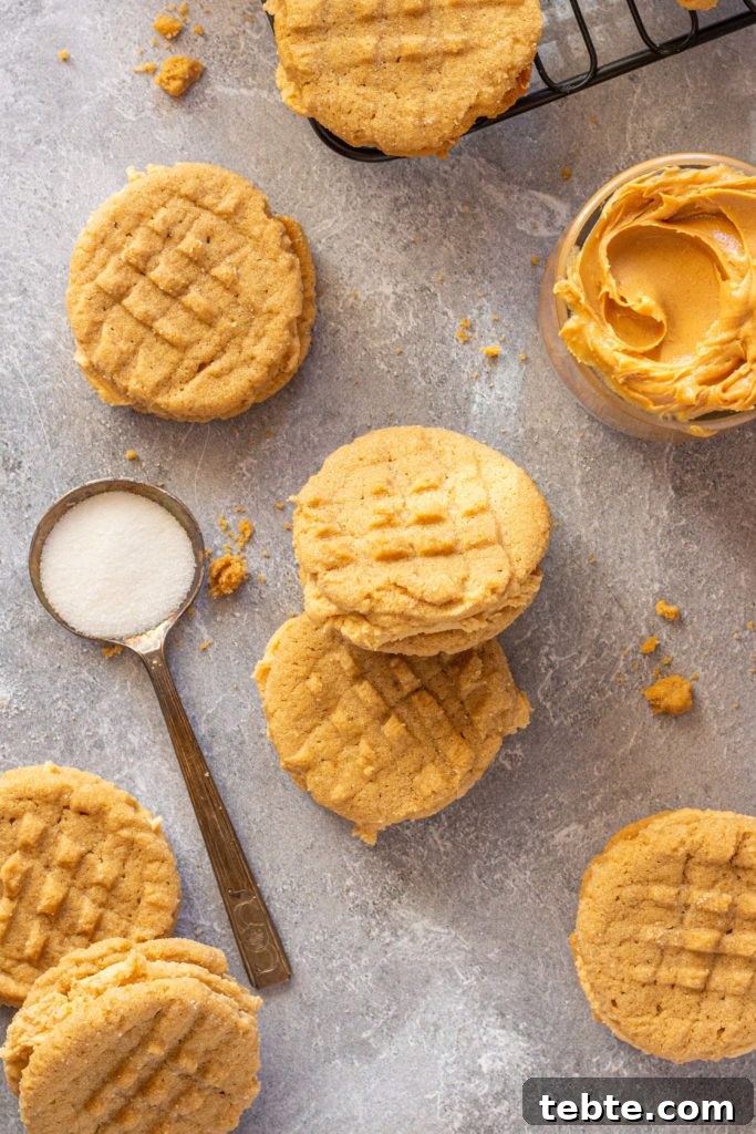 Gluten-Free Nutter Butter Cookies Recipe: Homemade Peanut Butter Sandwich Cookies 2 gluten free nutter butter sandwich cookies and peanut butter cookies are scattered on a gray marbled backdrop. a glass bowl of creamy peanut butter sits in the top right corner, and a spoon of white sugar is laid on the left side.