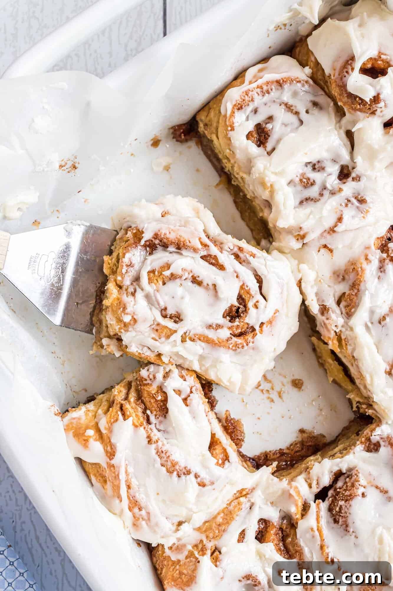Quick Crescent Roll Cinnamon Swirl Rolls 2 A spatula lifts a frosted cinnamon roll from a baking dish containing several iced cinnamon rolls.