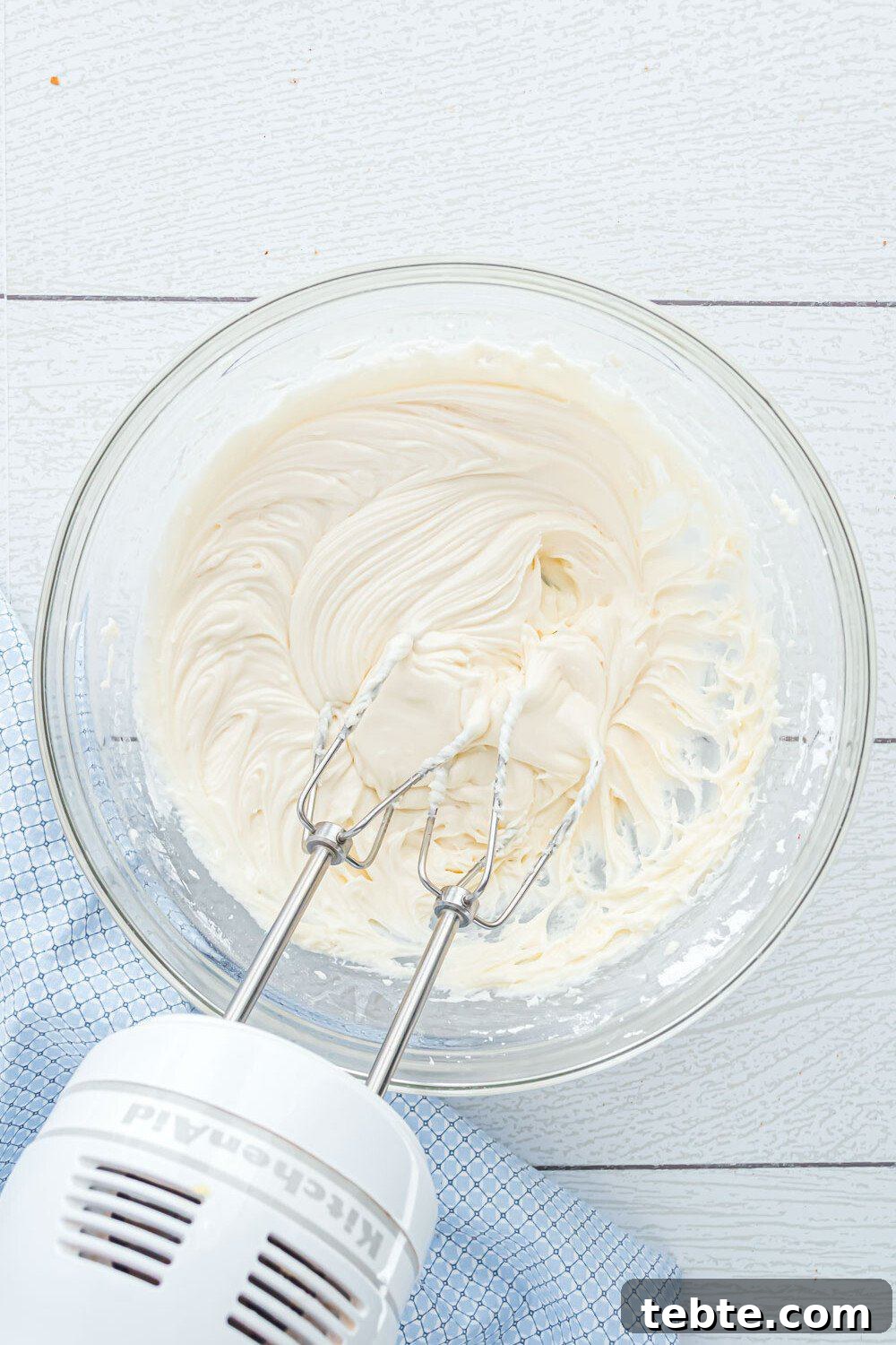 Quick Crescent Roll Cinnamon Swirl Rolls 4 A hand mixer with beaters is resting in a glass bowl of creamy, whipped mixture on a light wooden surface with a blue cloth nearby.