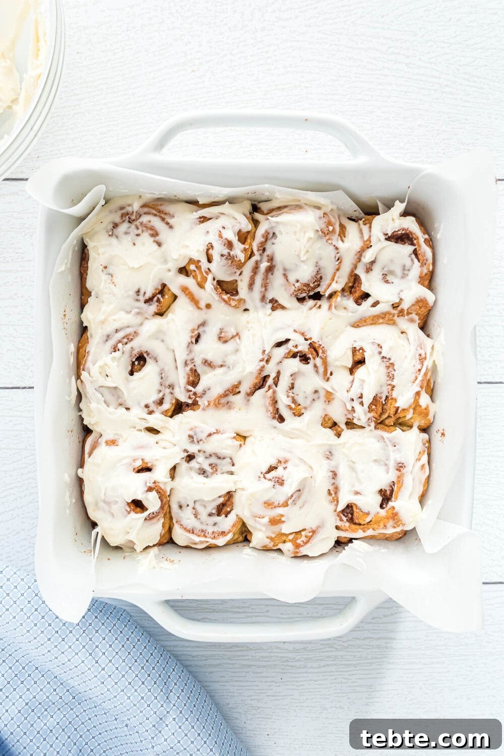 Quick Crescent Roll Cinnamon Swirl Rolls 6 A white baking dish filled with twelve cinnamon rolls topped with white icing, placed on a white surface next to a blue cloth.