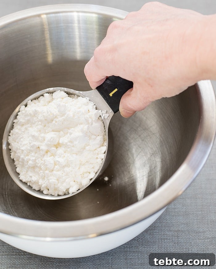 How to Make Natural DIY Body Powder at Home 3 A hand holding a measuring cup filled with cornstarch ready to put in a stainless mixing bowl