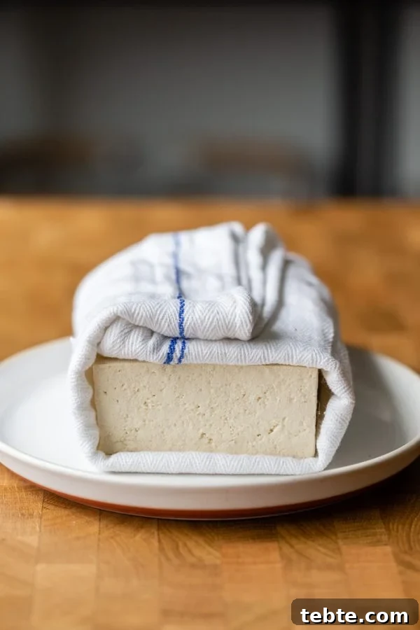 How to Press Tofu: Quick Methods With and Without a Tofu Press 4 block of tofu wrapped in a white and blue towel, sitting on a white plate. It's all on a butcher block counter