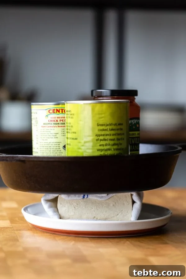 How to Press Tofu: Quick Methods With and Without a Tofu Press 5 block of tofu wrapped in a white and blue towel, sitting on a white plate. It's all on a butcher block counter. a skillet and heavy cans sitting on top to press the tofu