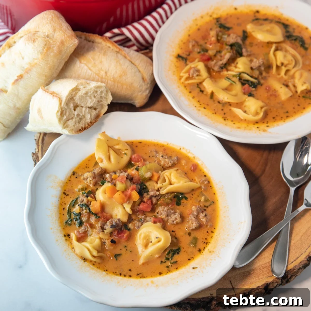 two bowls of spicy tortellini soup with bread and spoons sitting beside