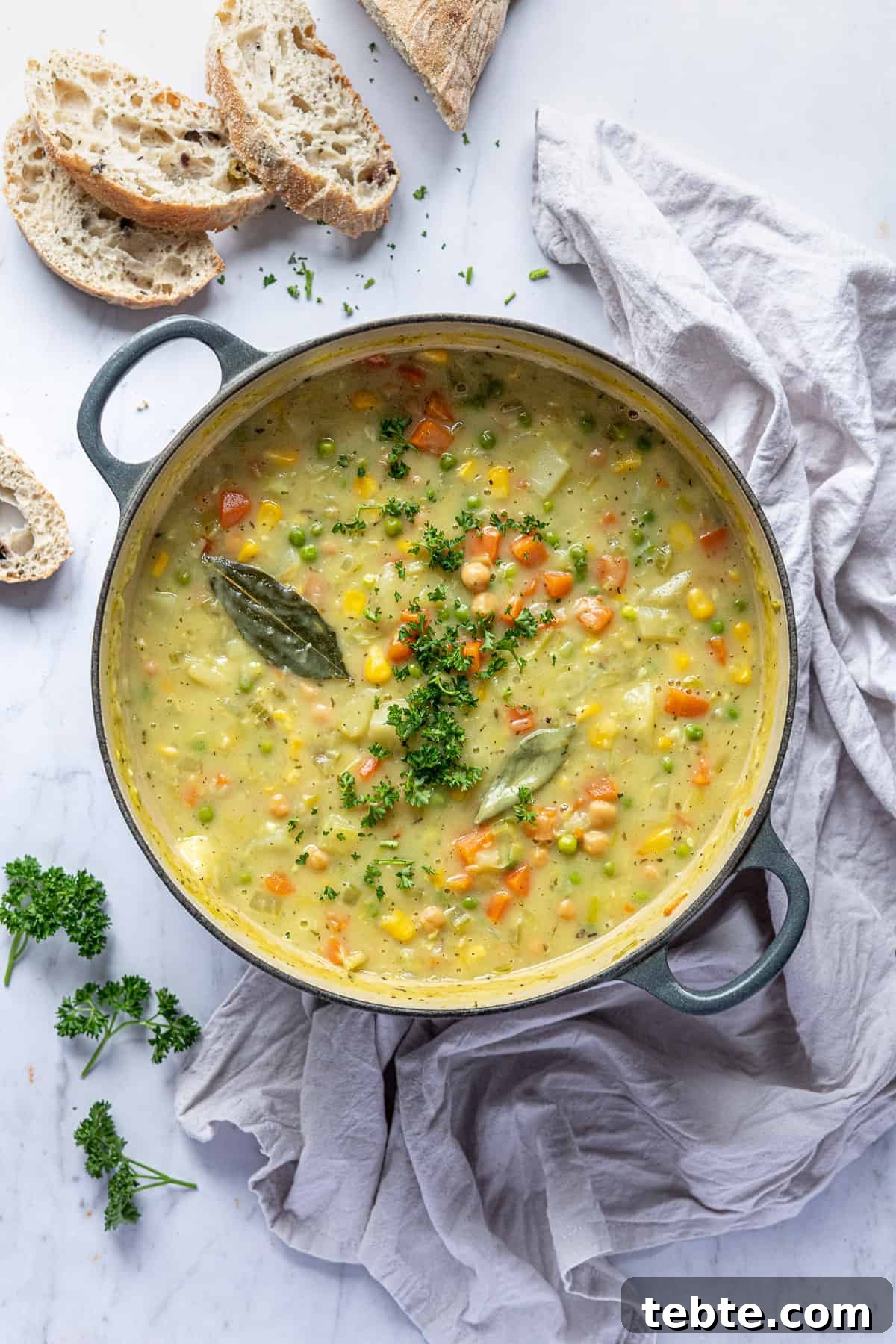 Creamy vegetable chickpea soup in a cast iron pan on a marble surface with sliced bread, fresh parsley and a grey cloth.