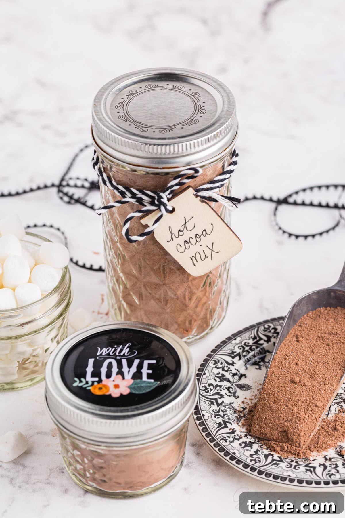 DIY Hot Cocoa Mix Recipe for Rich, Creamy Hot Chocolate 2 Two glass jars with homemade hot cocoa mix, decorative ribbon, and gift tags, glass jar with mini marshmallows, measuring cup with homemade hot cocoa mix, on a marble countertop.