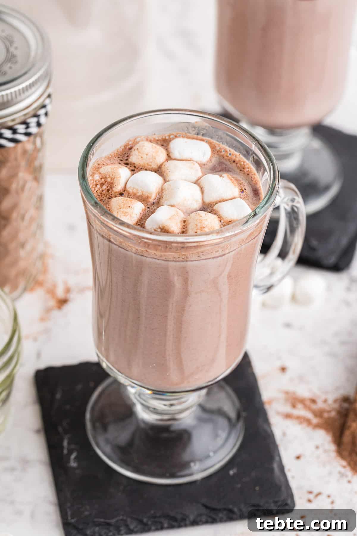 DIY Hot Cocoa Mix Recipe for Rich, Creamy Hot Chocolate 3 Two tall glasses with hot cocoa and mini marshmallow topping on top of black coasters, glass sealed jar filled with homemade hot cocoa mix, on a marble countertop.