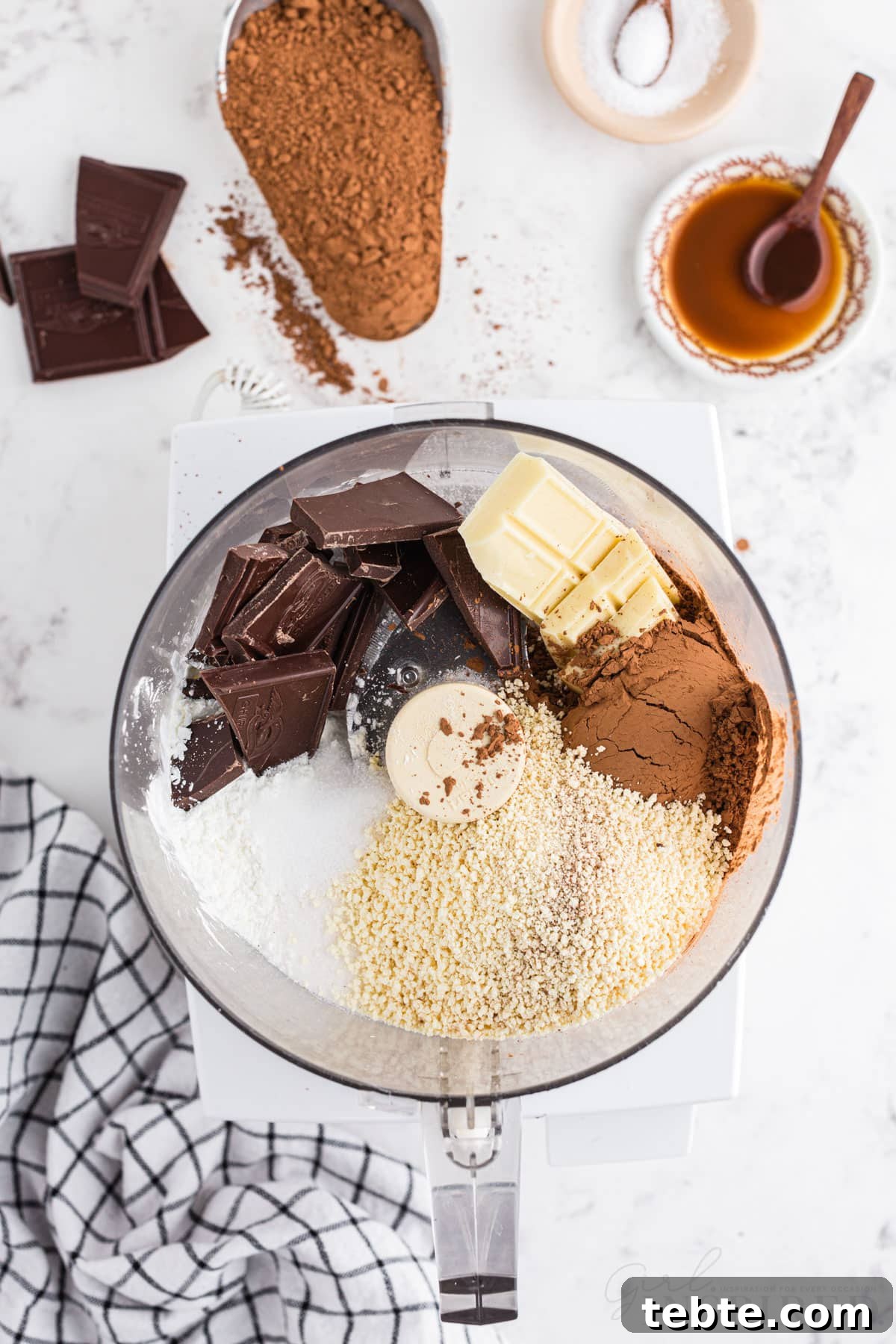 DIY Hot Cocoa Mix Recipe for Rich, Creamy Hot Chocolate 5 Hot cocoa ingredients added to a food processor, checkered linen, pieces of semi-sweet chocolate bar, metal measuring cup with unsweetened cocoa powder, bowl of sea salt, on a marble countertop.