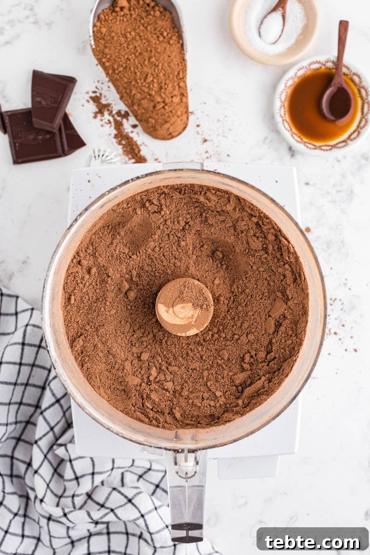 DIY Hot Cocoa Mix Recipe for Rich, Creamy Hot Chocolate 6 Blended homemade hot cocoa mix in a food processor, checkered linen, pieces of semi-sweet chocolate bar, unsweetened cocoa powder, sea salt, on a marble countertop.