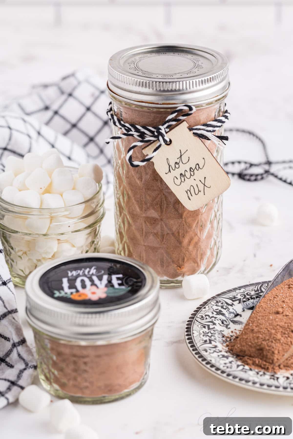 DIY Hot Cocoa Mix Recipe for Rich, Creamy Hot Chocolate 7 One large and one small glass jars filled with homemade hot cocoa mix with decorative ribbon and gift tags, glass jar with mini marshmallows, checkered linen, metal measuring cup with hot cocoa mix, on a marble countertop.