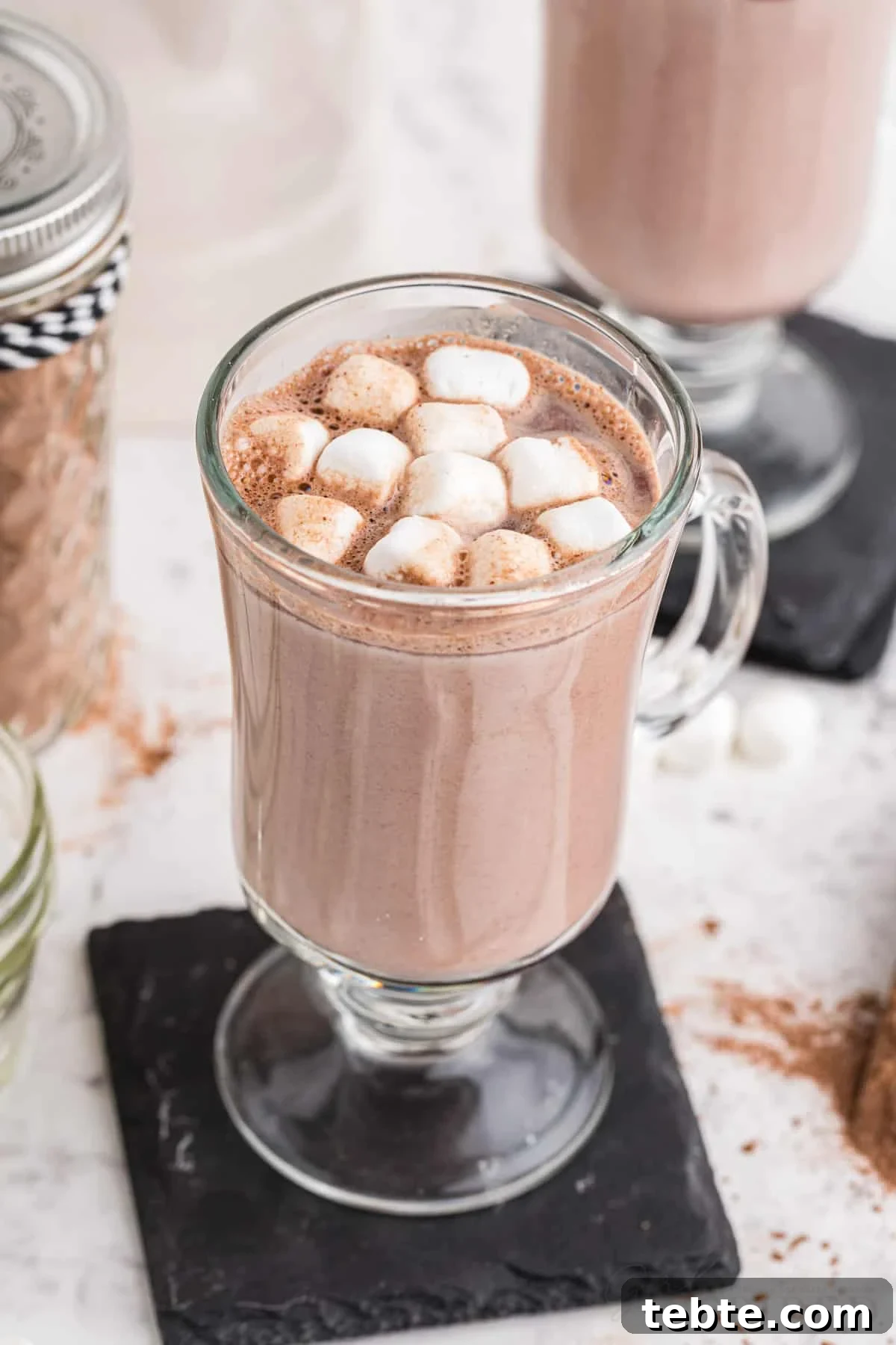 DIY Hot Cocoa Mix Recipe for Rich, Creamy Hot Chocolate 8 Two tall glasses with hot cocoa and mini marshmallow topping on top of black coasters, glass sealed jar filled with homemade hot cocoa mix, on a marble countertop.