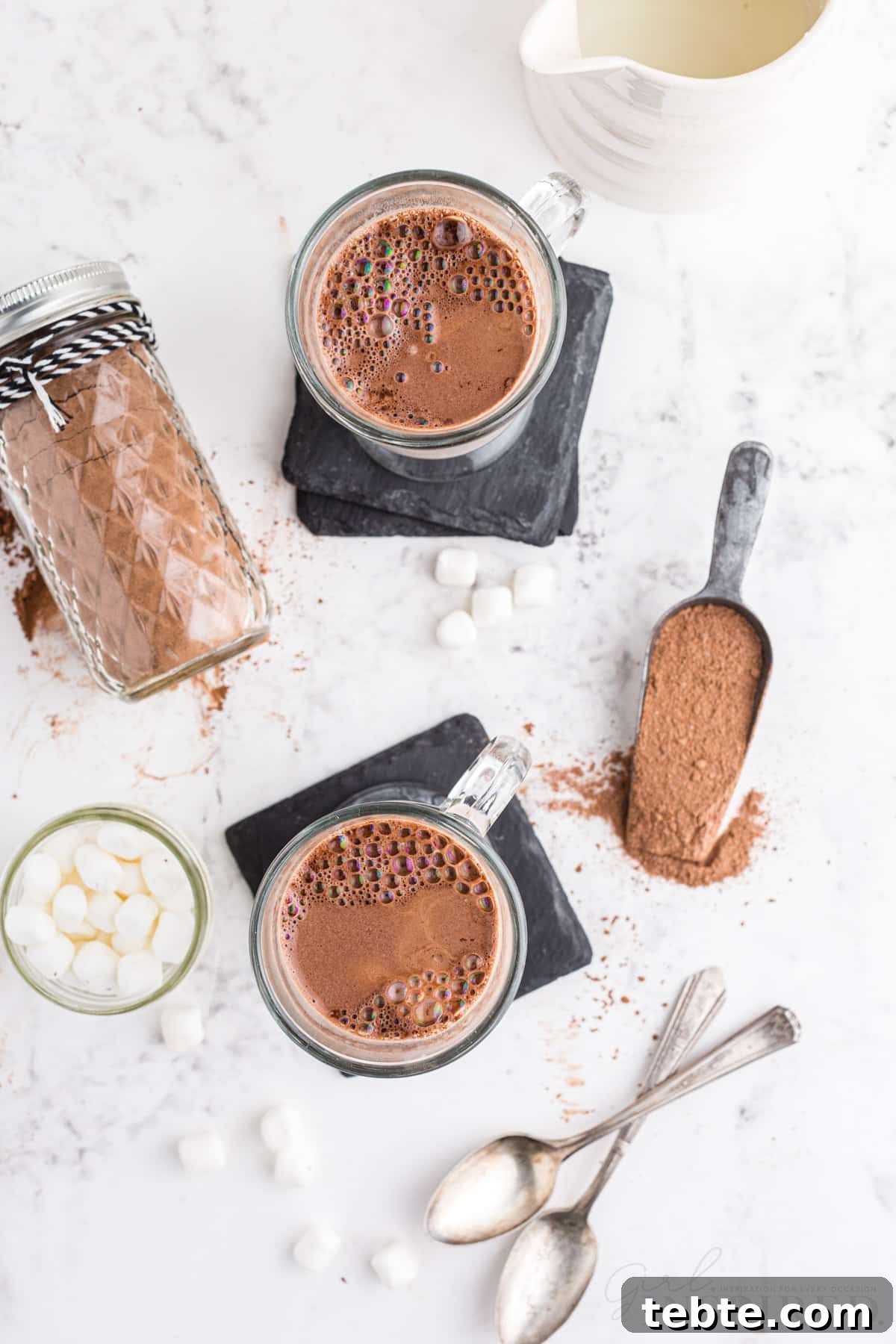 DIY Hot Cocoa Mix Recipe for Rich, Creamy Hot Chocolate 9 Overhead of two glasses of hot cocoa, two metal spoons, glass jar with mini marshmallows, jug with milk, metal measuring cup with hot cocoa mix, on a marble countertop.