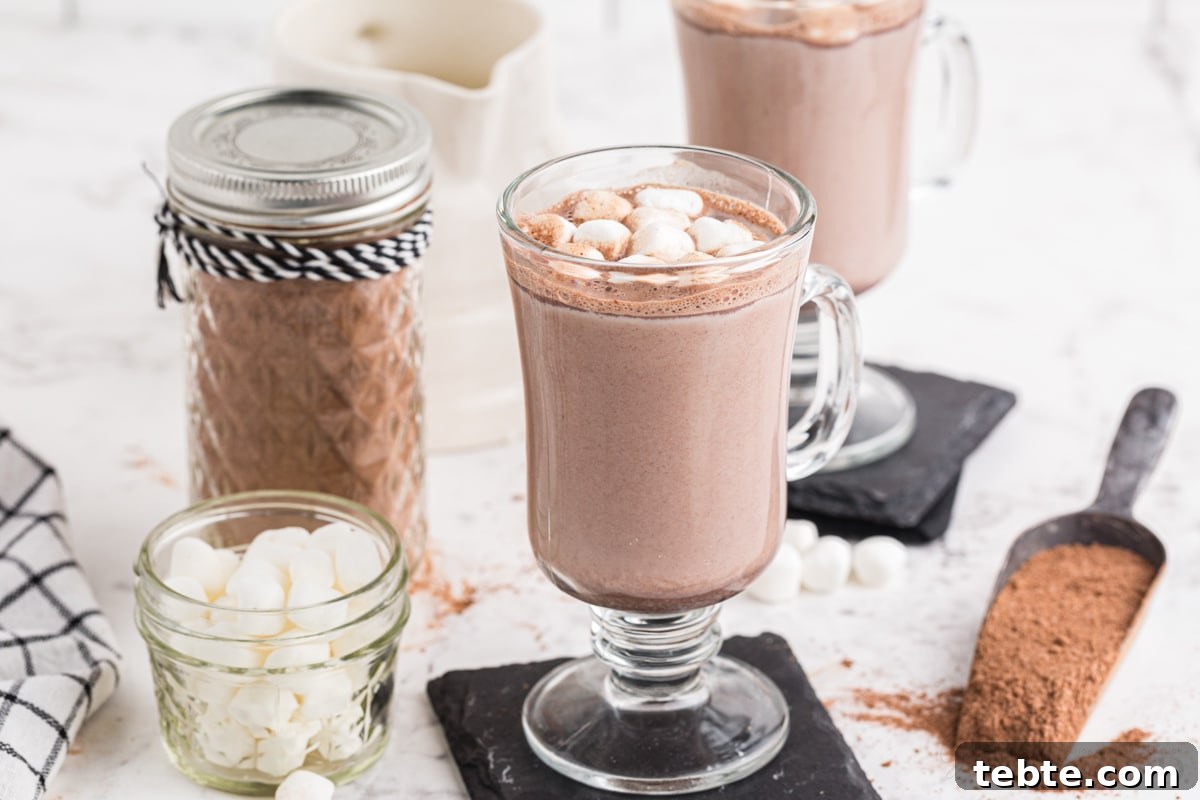 DIY Hot Cocoa Mix Recipe for Rich, Creamy Hot Chocolate 10 Two glasses of hot cocoa topped with mini marshmallows, glass jar with homemade hot cocoa mix with decorative ribbons, glass jar with mini marshmallows, checkered linen, metal cup with hot cocoa mix, on a marble countertop.