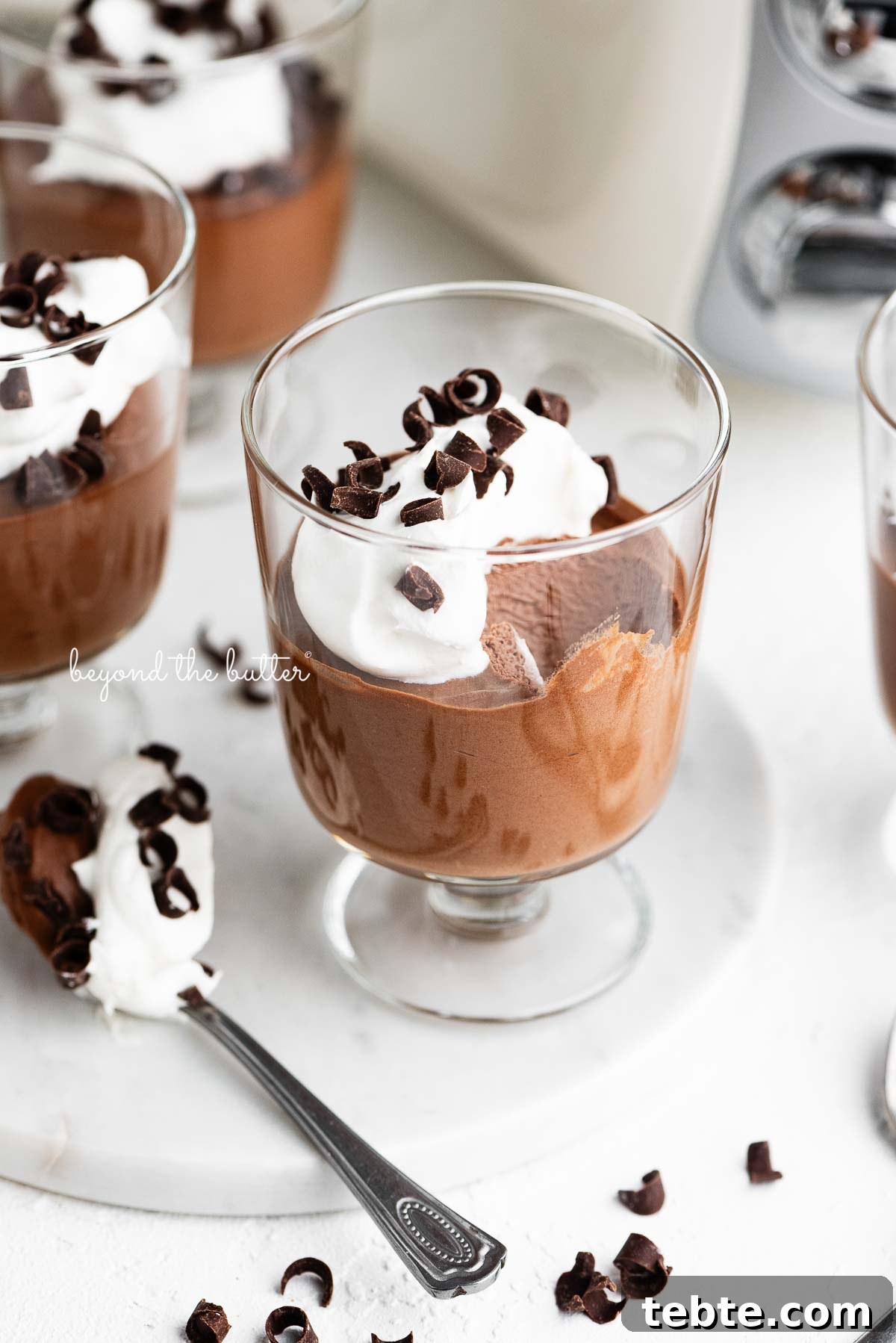Silky Blender Chocolate Mousse Recipe You Can Make in Minutes 2 Parfait goblets filled with chocolate mousse, whipped cream, and chocolate curls with a spoonful laying next to one on a white marble slab and white textured background.
