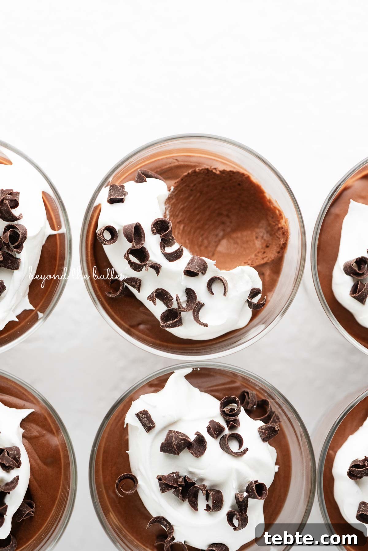 Silky Blender Chocolate Mousse Recipe You Can Make in Minutes 4 Six parfait goblets filled with chocolate mousse, whipped cream, and chocolate curls with a spoonful removed from the top center goblet on a white textured background.