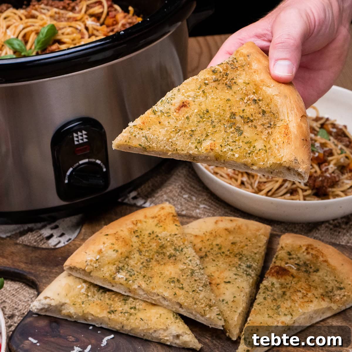 Crispy Garlic Pizza Bread Recipe — Cheesy Garlic Pull-Apart Loaf 35 Holding a slice of Garlic Pizza Bread over a bowl of spaghetti bolognese and a slow cooker in the back ground