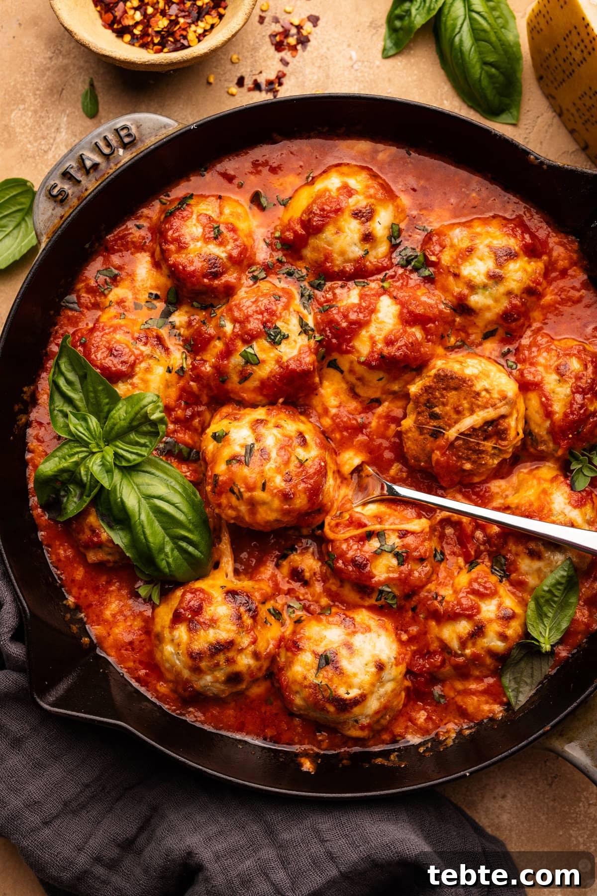 Chicken Parmesan Meatballs with Zucchini Skillet 2 A skillet pan with chicken parmesan meatballs in it with fresh basil and a spoon in the pan. The skillet is sitting on a tan background with a herbs, a block of cheese, and a small bowl of red pepper flakes placed around it.
