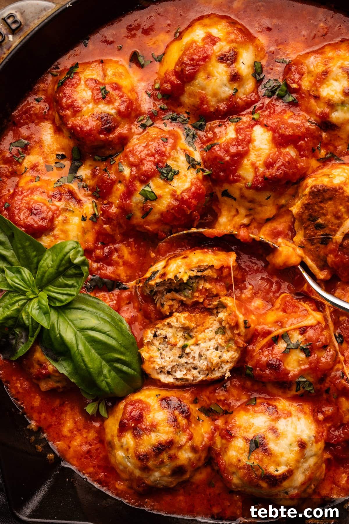 Chicken Parmesan Meatballs with Zucchini Skillet 6 A close up of chicken parmesan meatballs, one of the meatballs is cut in half with a spoon and fresh basil next to it.