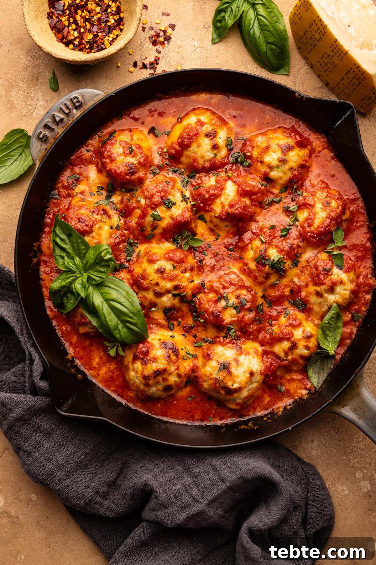 Chicken Parmesan Meatballs with Zucchini Skillet 7 A skillet pan with chicken parmesan meatballs baked in it with fresh basil on top. The pan is sitting on a tan background with basil leaves, a block of cheese, and a small bowl arranged around it.