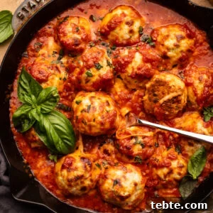Chicken Parmesan Meatballs with Zucchini Skillet 8 A skillet pan with chicken parmesan meatballs in it with fresh basil and a spoon in the pan. The skillet is sitting on a tan background with a herbs, a block of cheese, and a small bowl of red pepper flakes placed around it.
