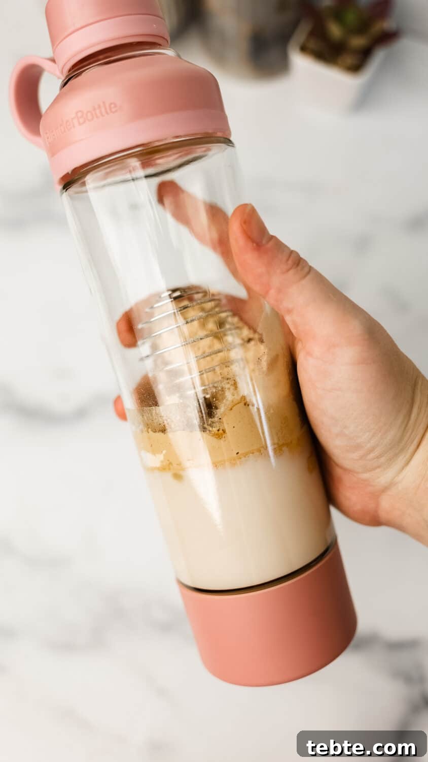 Hand holding a pink blender bottle filled with almond milk, protein powder, and spices before shaking.