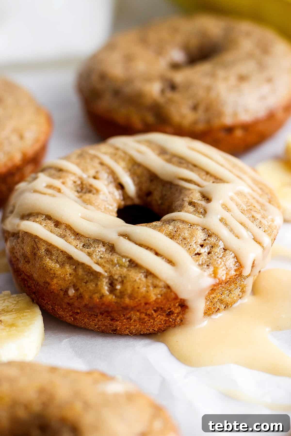 A close-up image of a glazed banana doughnut, with more doughnuts and banana slices in the background.