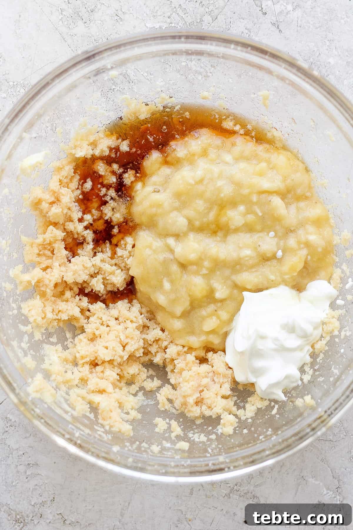 Ingredients combined in a glass bowl, possibly for baking, including mashed bananas, sugars, and a dollop of sour cream or yogurt.