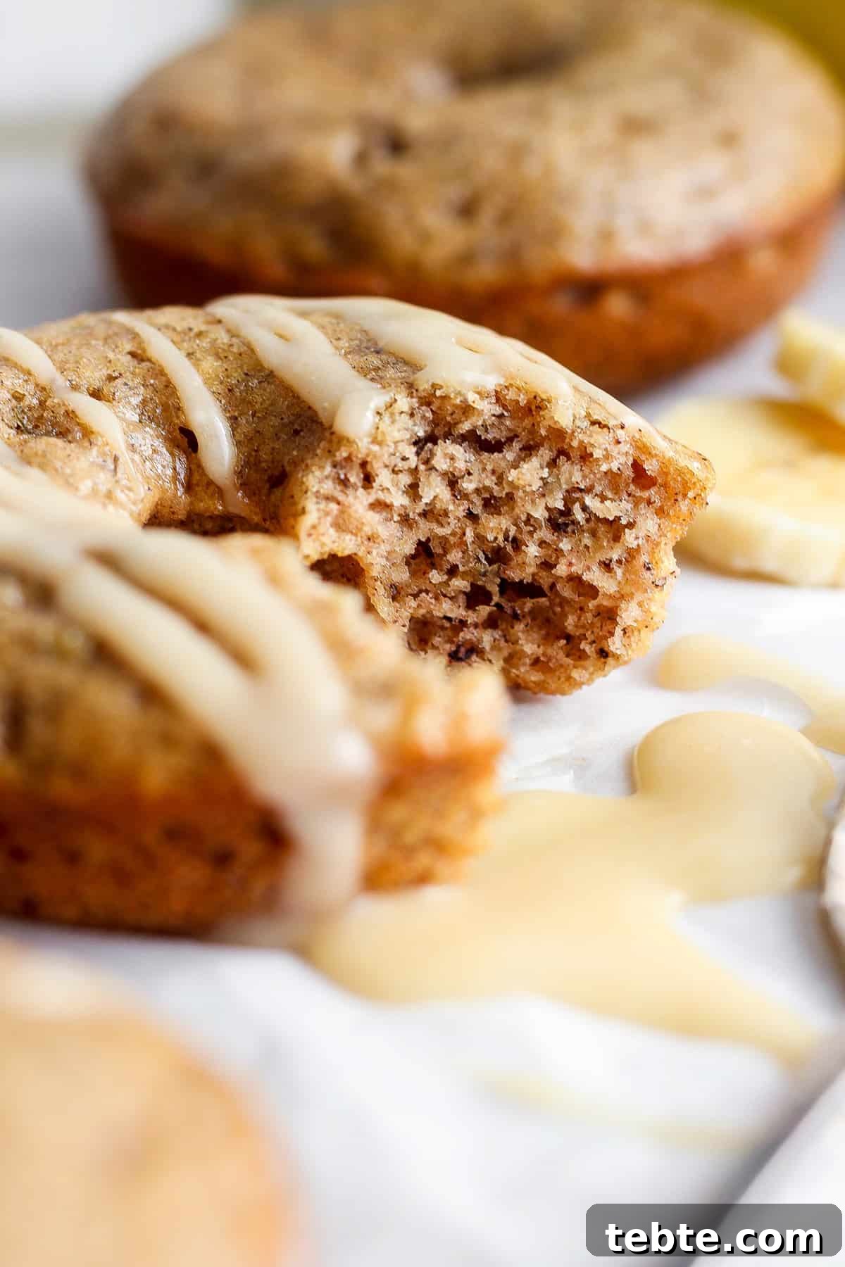 Close-up of a partially eaten muffin with white glaze on top, with more muffins and banana slices blurred in the background.