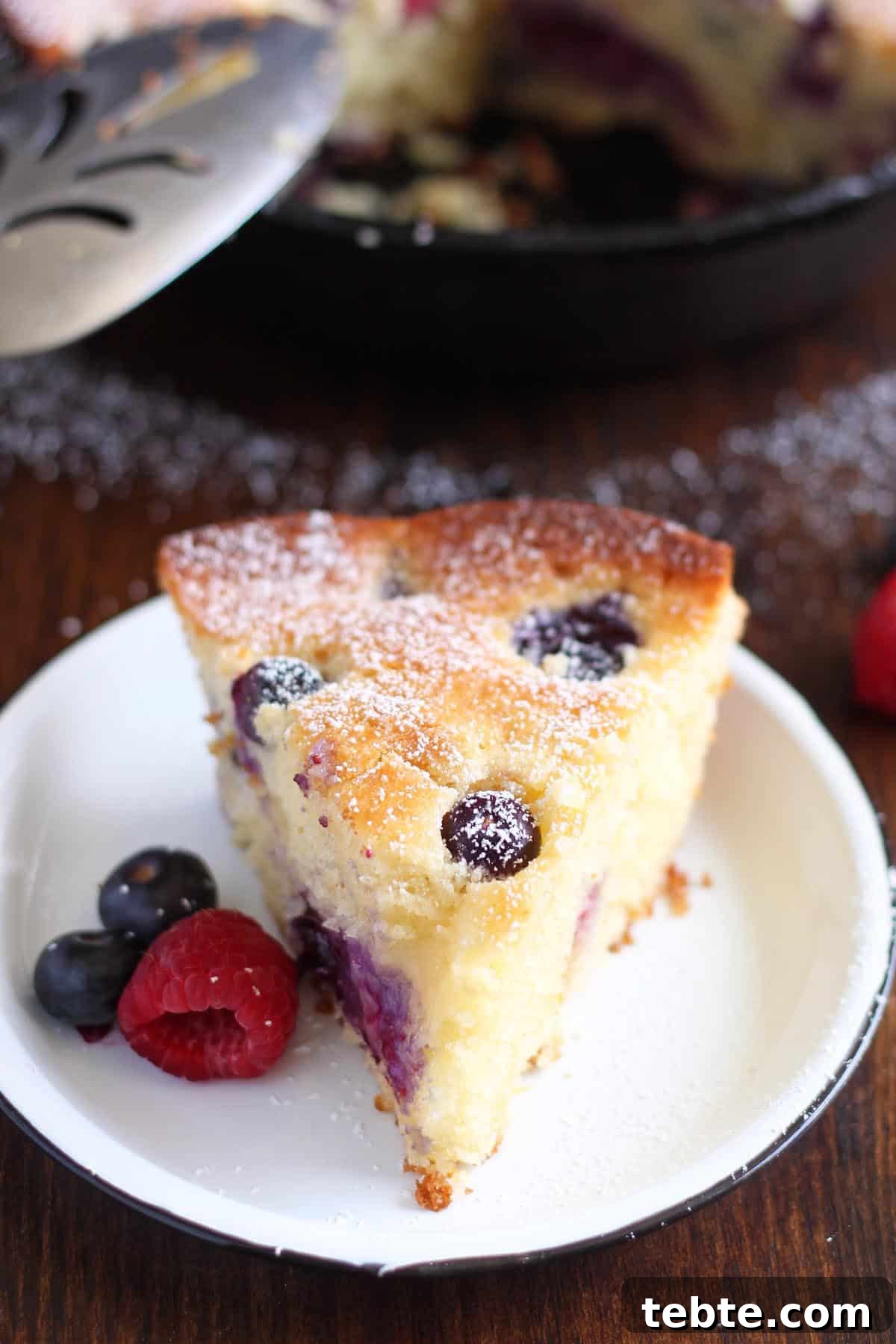 This Mascarpone Mixed Berry Cake is extremely soft and moist, bake it and serve it straight from the pan. It’s excellent as a coffee cake for breakfast or as dessert with a scoop of ice cream on the side. #wildwildwhisk #berrycake 3/4 view: a slice of mascarpone cake on a white plate next to some fresh berries.
