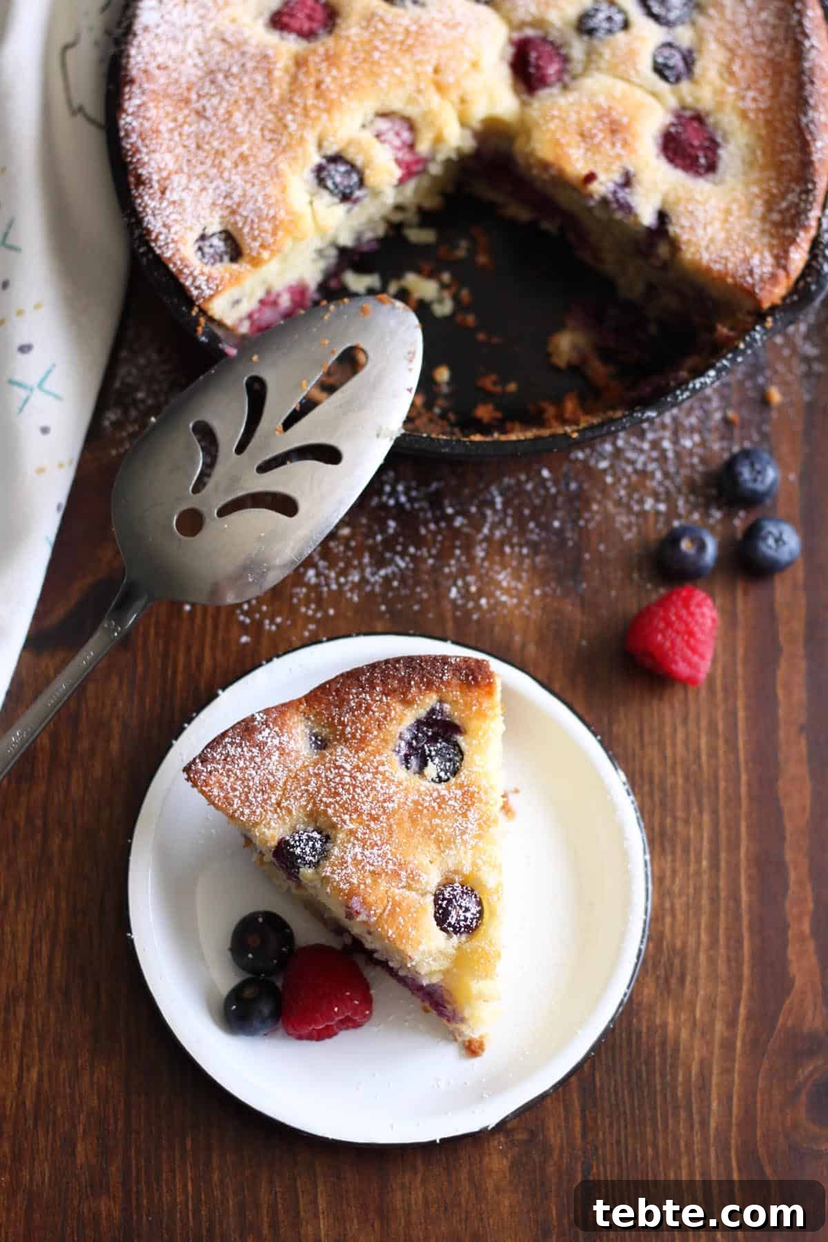 This Mascarpone Mixed Berry Cake is extremely soft and moist, bake it and serve it straight from the pan. It’s excellent as a coffee cake for breakfast or as dessert with a scoop of ice cream on the side. #wildwildwhisk #berrycake A slice of mascarpone berry cake on a plate next to the cast iron skillet that the cake was baked in.