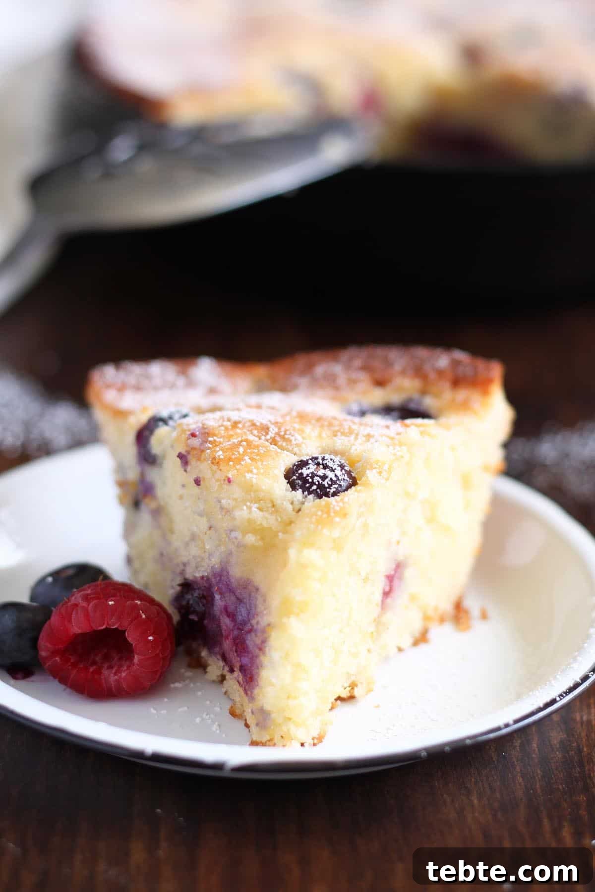 This Mascarpone Mixed Berry Cake is extremely soft and moist, bake it and serve it straight from the pan. It’s excellent as a coffee cake for breakfast or as dessert with a scoop of ice cream on the side. #wildwildwhisk #berrycake Angle view: a slice of mascarpone mixed berry cake on a white plate next to some fresh berries.