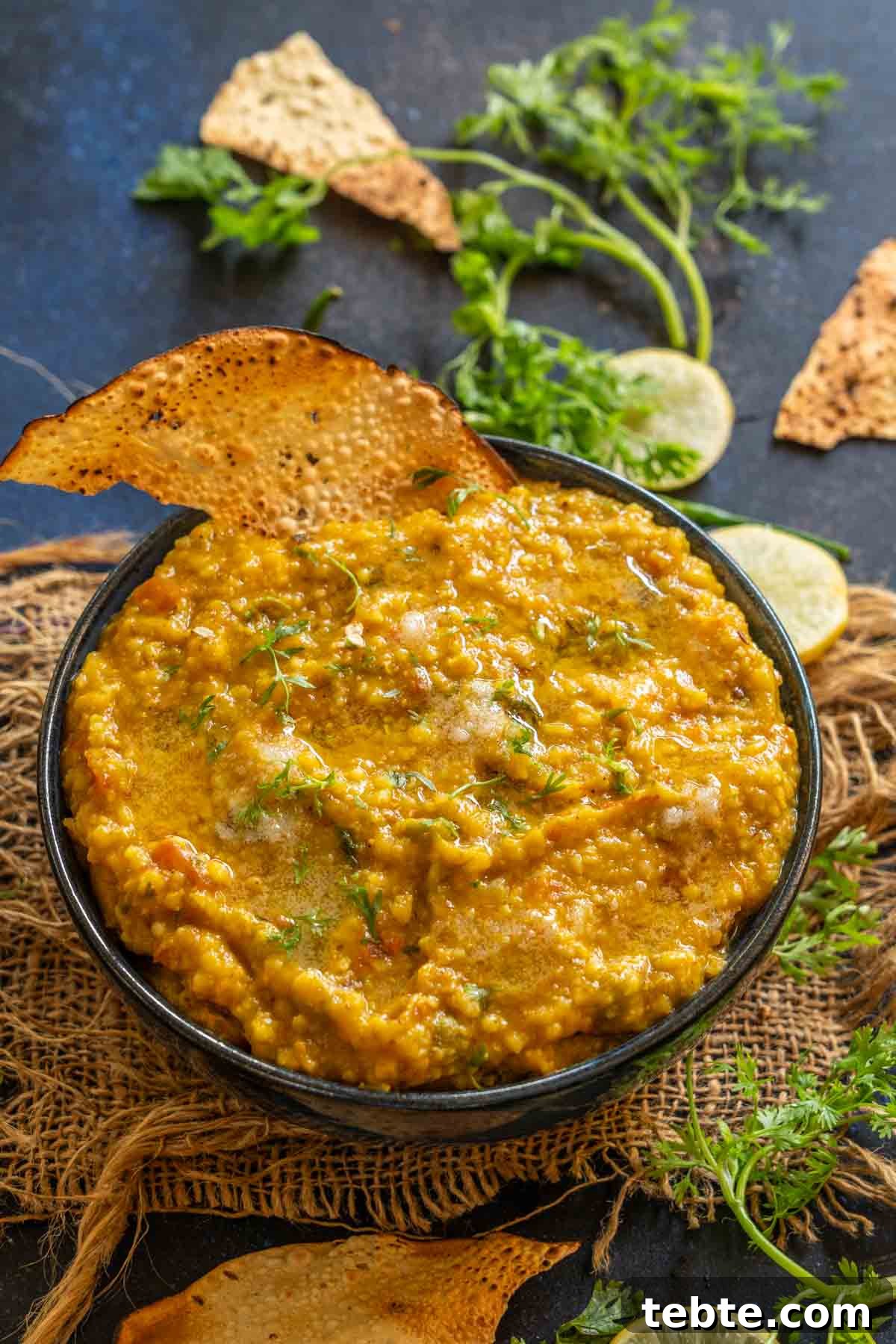 Jowar Khichdi Recipe: Wholesome Millet One-Pot Meal 2 A bowl of jowar khichdi garnished with fresh herbs, served alongside crispy papad and slices of lemon, chopped cilantro, and broken papad are scattered on a dark surface in the background.