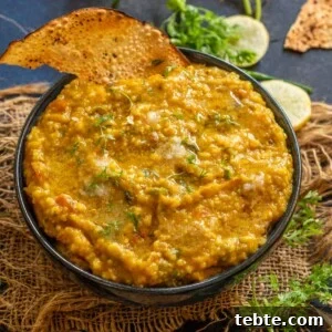 Jowar Khichdi Recipe: Wholesome Millet One-Pot Meal 13 A bowl of jowar khichdi garnished with fresh herbs, served alongside crispy papad and slices of lemon, chopped cilantro, and broken papad are scattered on a dark surface in the background.