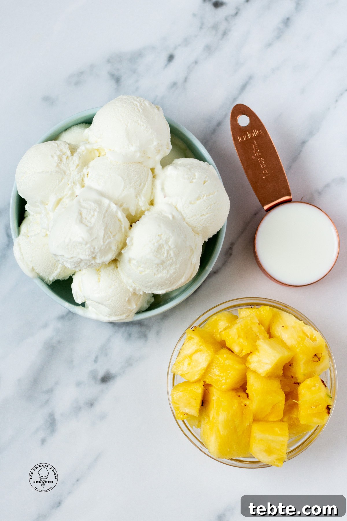 Creamy Pineapple Milkshake Recipe – Tropical Smoothie Treat 3 a large bowl of scooped vanilla ice cream, a smaller bowl of diced pineapple, and a measuring cup holding milk, on a marble counter, viewed from above.
