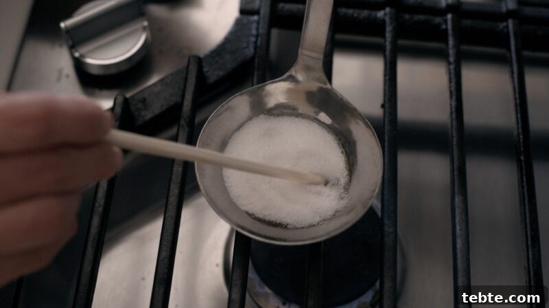5-Minute Dalgona Candy Recipe: Crispy Korean Sugar Treat 7 A wooden chopstick is stirring white sugar in a ladle over low heat on the stove.