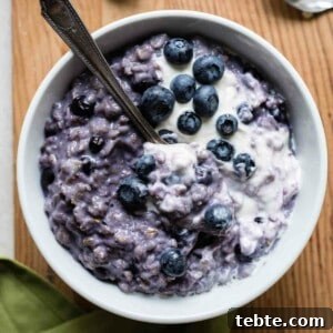Bowl filled with purple colored blueberry oatmeal, topped with cream and fresh blueberries.